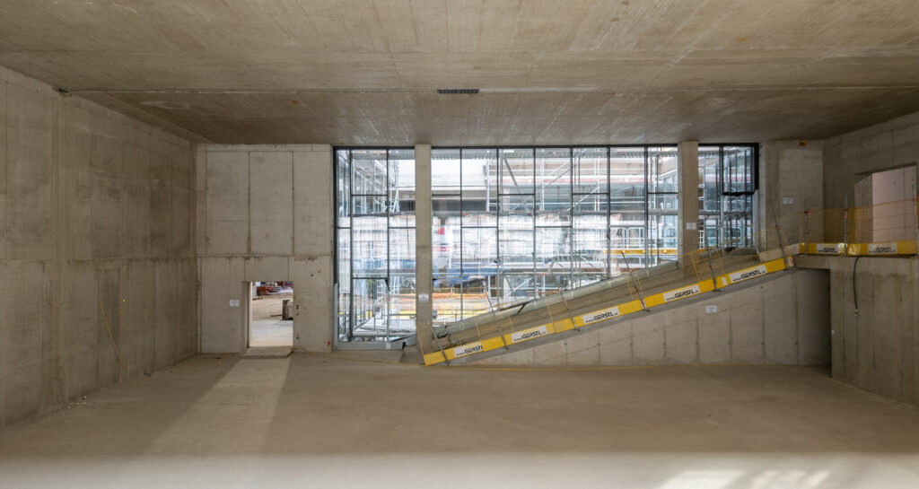 Baustellenfoto eines Hörsaals. Es ist viel Beton zu sehen, und die Glasfassade ist bereits erkennbar.