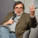 Ein sitzender Mann spricht ins Mikrofon und hebt die Hand.