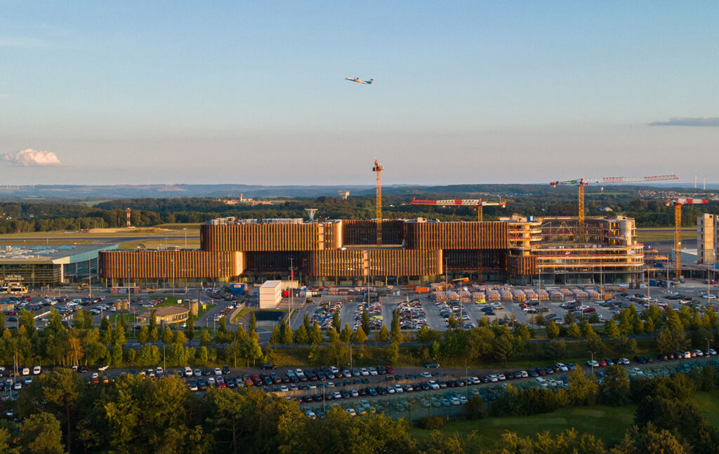 Statt eines einzigen, langgezogenen Körpers mäandert das aus zwei jeweils dreigeschossigen Baukörpern bestehende Gebäude auf einer Länge von 365 Metern entlang des Flugfelds. © Weidart.lu