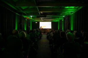 Ein dunkler Raum mit grüner Beleuchtung, einer Bühne und vielen Zuschauer*innen.