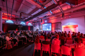 Ein rot ausgeleuchteter Raum mit vielen Stühlen die zu einer Bühne ausgerichtet sind. Die Besucher*innen lauschen einen Vortrag.