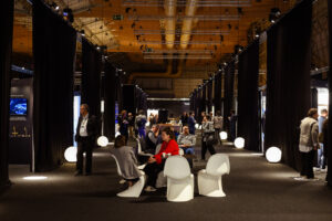 Besucher*innen sitzen auf weißen Designersesseln in einer dunklen Halle.