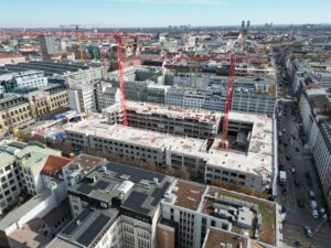 Luftaufnahme der Baustelle von The Verse: Im Herzen der Münchner Innenstadt entsteht auf sechs Vollgeschossen und zwei Staffelgeschossen ein Mixed-Use-Quartier mit über 70.000 Quadratmetern Mietfläche und Raum für mehr als 3.500 Arbeitsplätze.© Oaktree / Accumulata