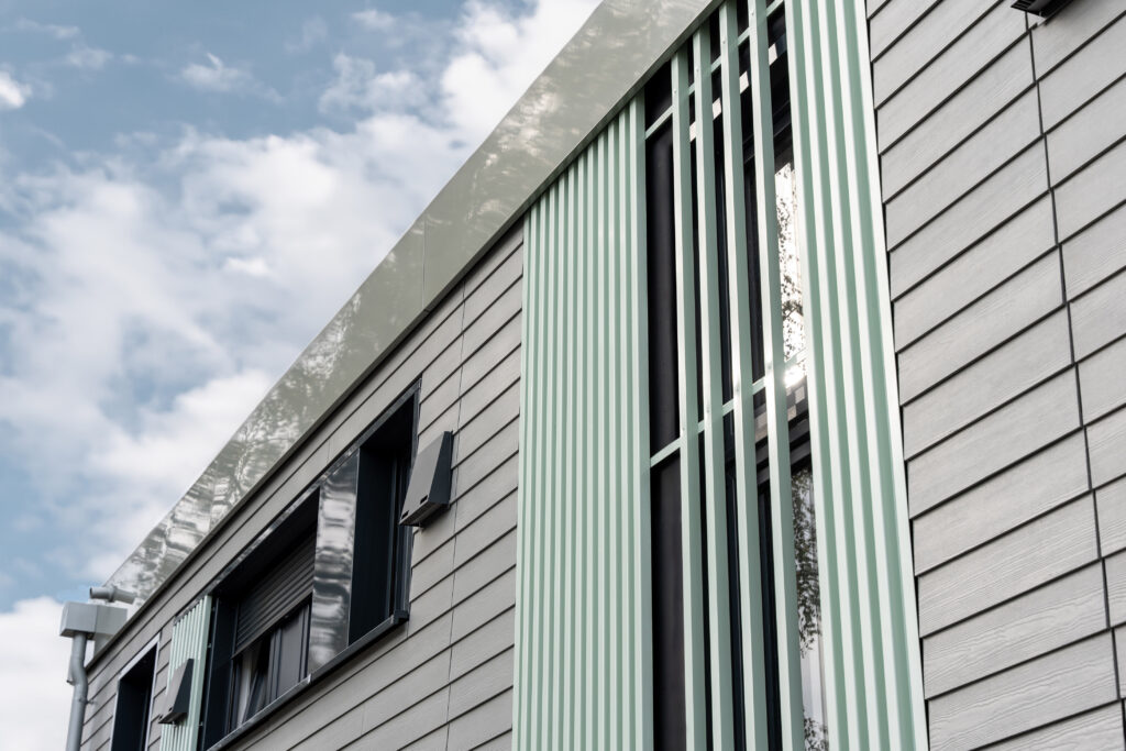  Die grauen Fassadenmodule sind mit Faserzement-Platten in Holzoptik bekleidet und mit Zel-lulose- oder Holzfaser gedämmt. Lisenen werten die Fassade auf und kaschieren das für die Vorfertigung charakteristische Fugenbild. © Ecoworks