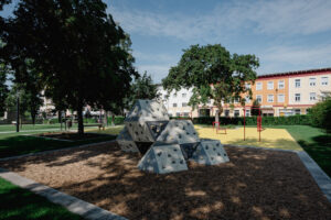 Ein architektonisch gestalteter Grünbereich mit Bäumen und einer Skulptur. Im Hintergrund ein Haus.