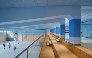 Ein Innenraum einer Schule. Eine Galerie mit hellem Holzböden und Blauen Säulen lässt Besucher*innen ins untere Geschoss blicken.