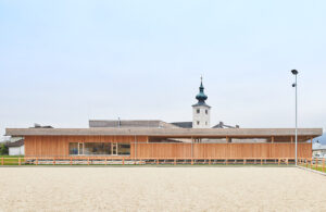 Ein flaches, langes Gebäude vor einer Kirche. Davor ist ein großer leerer Platz.