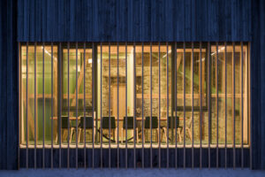 Ein Glaskubus in der Nacht. Er ist von innen beleuchtet, man kann Sessel um einen Tisch erkennen.