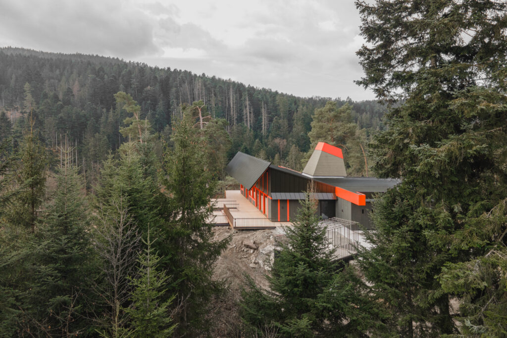 Ein Gebäude steht mitten im Wald. Es hat rote Dachelemente und schaut modern aus.