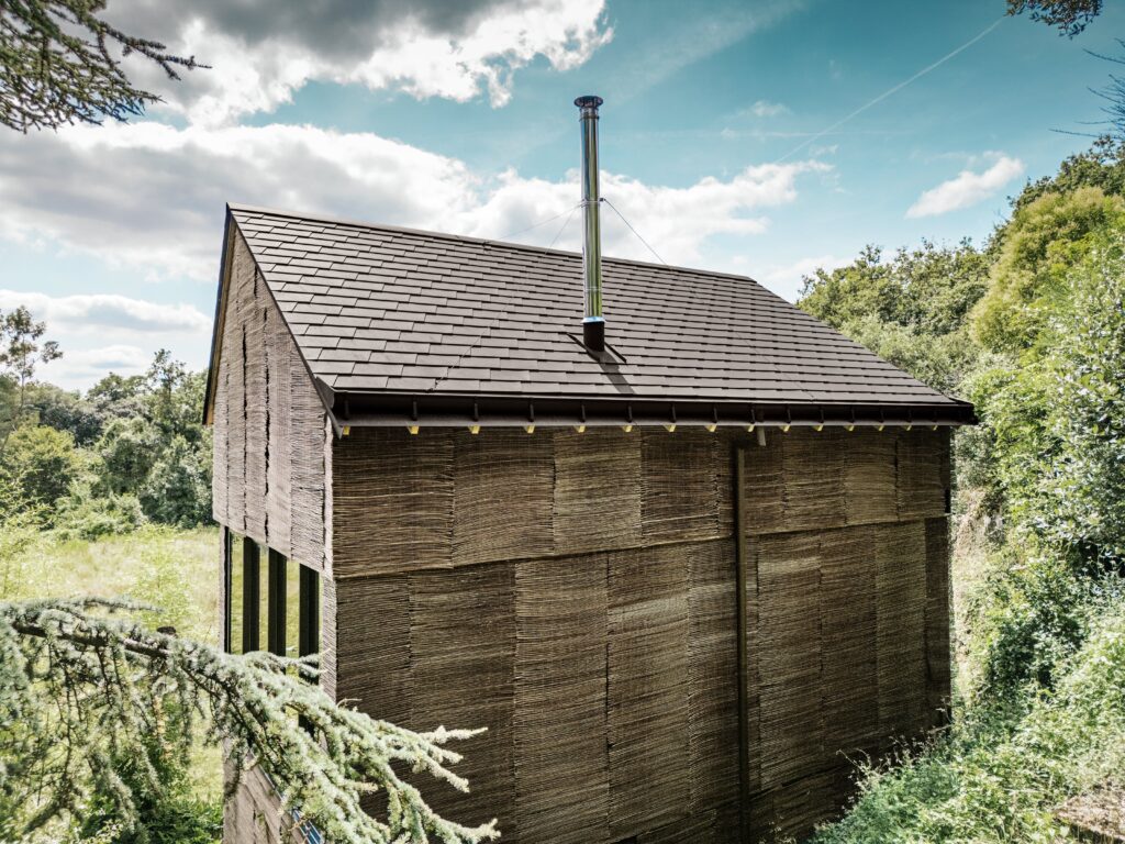 Die ruhige, materialgeprägte Erscheinung des Hauses fügt sich harmonisch in die bewaldete Umgebung ein. Die Fassade ist mit Weidenmatten bekleidet, die über eine schwarze Dichtfolie montiert wurden.