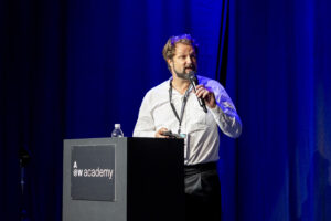 Ein Mann im Anzug hält einen Vortrag auf einer Bühne vor einem blauen Vorhang.