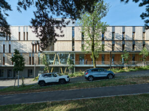 Holzfassade am Hang eines Hügels zu sehen. Davor parken Autos.